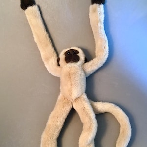 May include: A plush monkey toy with long, cream-colored limbs and a brown face, hands, and feet. The monkey's arms are raised, and its tail is curved. The toy is made of soft, fuzzy material.