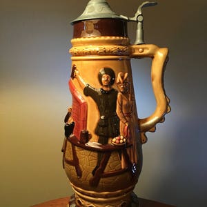 May include: A tall, decorative beer stein with a hinged metal lid and handle. The ceramic body features a relief design of figures in medieval attire, with brown, tan, and gold tones. The base has a decorative, scalloped edge.