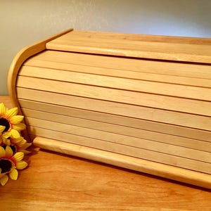 May include: A light-colored wooden bread box featuring a roll-top tambour door design with horizontal slats. The rectangular container has curved side panels and a smooth finish, positioned next to a cluster of yellow artificial sunflowers.