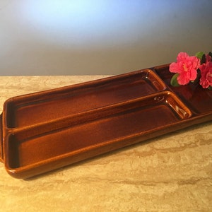 May include: A rectangular, brown ceramic serving tray with three compartments. Two long, narrow sections are separated by a smaller square section. Two pink flowers rest on the tray. The tray has handles on each end.