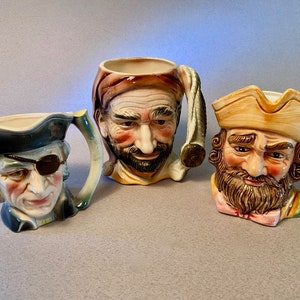 May include: Three ceramic mugs with detailed, hand-painted faces of historical figures. One mug features a pirate with an eye patch, another a man with a turban, and the third a bearded man in a tricorn hat. Each mug has a handle and is painted in vibrant colors.