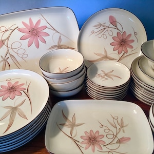 May include: A vintage cream-colored dinnerware set featuring a floral design with pink flowers and brown accents. The set includes plates, bowls, saucers, and cups. The edges of the dishes have a brown trim.