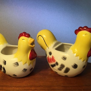 May include: Two ceramic chicken-shaped creamers. The creamers are yellow with red combs and beaks, and white bodies with black and red markings. One creamer has an open beak, and the other has a closed beak.
