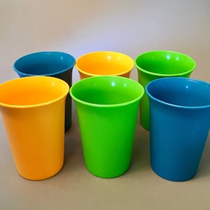 May include: Six colorful plastic cups in shades of yellow, green, and blue are arranged on a neutral surface. The cups have a slightly tapered design, with a wider opening at the top. The colors are vibrant and evenly distributed.