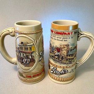 May include: Two beige ceramic beer steins with ornate handles. One stein features a building scene with a horse-drawn carriage and the text "THE STROH BREWERY". The other stein has a village scene and the text "STROH'S BREWERY COMPANY".