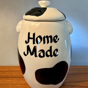 May include: A ceramic cookie jar with a lid, featuring a white base with black cow-like spots. The words "Home Made" are written in black script on the front. The jar has a handle on the lid and two small handles on the sides.