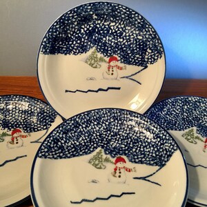 May include: Set of four white ceramic plates with a winter scene. Each plate features a snowman wearing a red hat and scarf, with a blue dotted border and a snowy landscape design.