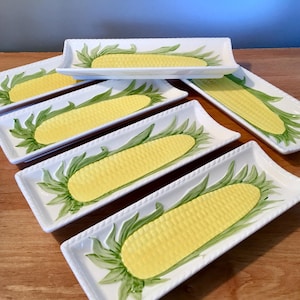 May include: A set of six rectangular white ceramic serving dishes. Each dish features a yellow corn cob design with green leaves. The edges of the dishes have a rope-like texture.