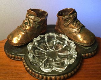 May include: A decorative ashtray featuring two bronze-colored baby shoe sculptures and a clear glass ashtray. The shoes are detailed with laces and sit on a dark metal base with a beaded edge. The glass ashtray has a floral design.