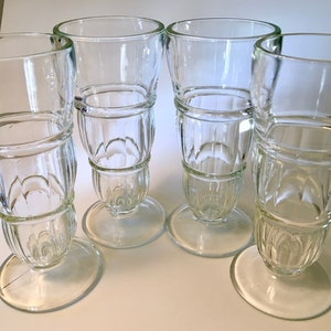 May include: Four clear, vintage-style drinking glasses. Each glass features a round base, a stem with a decorative, ribbed design, and a wide, cylindrical top. The glasses are arranged on a white surface.