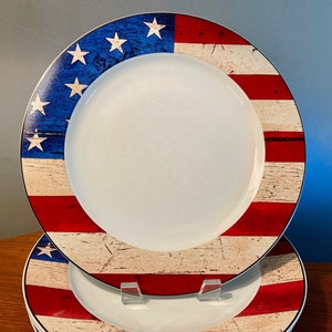 May include: White ceramic plates with a patriotic design. The rim features the American flag with red and white stripes, blue field with white stars, and a distressed wood-like texture. The plates are stacked on a wooden surface.
