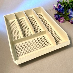 May include: Off-white plastic utensil tray with multiple compartments and a perforated base. The tray has a rectangular shape with raised sides and dividers to organize items. The design includes a series of small square holes throughout the base.
