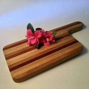 May include: A wooden cutting board with a handle, featuring alternating stripes of light and dark wood. A cluster of pink flowers rests on the board. The board is rectangular with rounded corners and a handle.