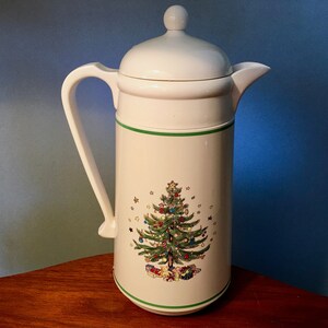 May include: A white thermal carafe with a handle, a spout, and a lid featuring a knob. The body is decorated with a green horizontal stripe at the top and bottom, and a central illustration of a decorated Christmas tree with gifts underneath.