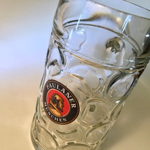 May include: Clear glass beer stein with a textured pattern and a circular logo that reads "PAULANER MUNCHEN" in black and gold lettering. The logo features a profile of a man in a brown robe against a red background.