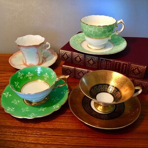 May include: Four antique teacups and saucers in various designs. One cup is green with white floral patterns, another is gold with black accents, and a third is light green with pink trim. The teacups are on a wooden surface.