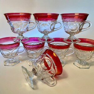 May include: A set of eight clear glass teacups with a ruby red rim and handle. The cups have a decorative pattern and are set on a pedestal base. One cup is lying on its side.