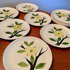 May include: A set of eight white ceramic plates. Each plate features a hand-painted design of a yellow flower with green leaves and a black branch. The plates are arranged on a wooden surface.