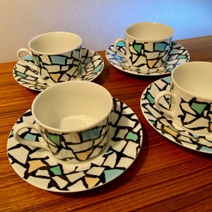 May include: Four white ceramic teacups and saucers. Each cup and saucer features a mosaic-like pattern in black, light blue, yellow, and green. The cups have a simple handle and a white interior. The set is displayed on a wooden surface.