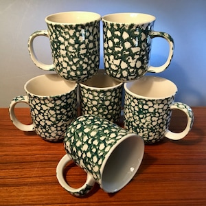 May include: A set of six ceramic mugs with a speckled green and white pattern. Each mug has a rounded body, a white interior, and a curved handle. The mugs are stacked on a wooden surface.