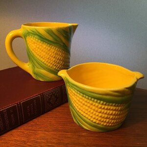 May include: A yellow and green ceramic pitcher and bowl set. The pitcher has a handle and spout, and the bowl has two small handles. Both pieces are decorated with a corn-on-the-cob design.