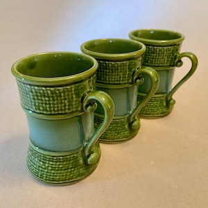 May include: Three green ceramic mugs with textured bands and curved handles. The mugs have a glossy finish and a slightly flared base. The design includes a woven pattern around the top and bottom of the mugs.