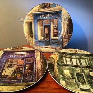 May include: Three decorative plates featuring Parisian storefronts. The plates depict a bookstore with the word "Livres", a shop named "Emilie", and a restaurant called "Chez Jerome". Each plate has a different color scheme, with blue, purple, and green storefronts.