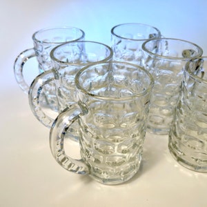 May include: Set of clear glass beer mugs with textured sides and sturdy handles. The mugs are arranged on a white surface, showcasing their classic design. The image highlights the transparency and the traditional shape of the glassware.