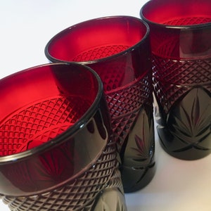 May include: Four ruby red drinking glasses with diamond and leaf pattern detailing. The glasses have a deep red color and are made of a thick, possibly crystal, material. The glasses are empty and are set against a white background.