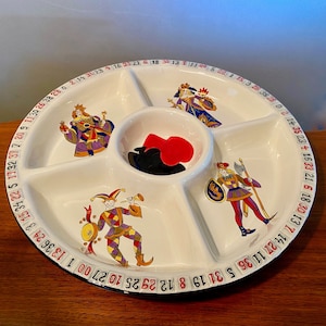 May include: White ceramic serving platter with five sections, featuring colorful illustrations of jesters and royalty. The platter has a black rim with numbers around the edge and contains red and black game pieces in the center.