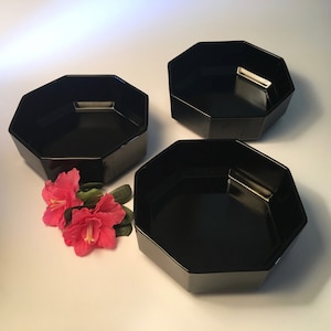 May include: Three black, octagonal serving bowls are arranged on a white surface. The bowls have a glossy finish and are empty. Pink flowers with green leaves are placed near one of the bowls.