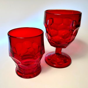 May include: Two ruby red drinking glasses. One is a short tumbler, and the other is a stemmed goblet. Both glasses have a textured, geometric pattern. The glasses are made of a transparent material.