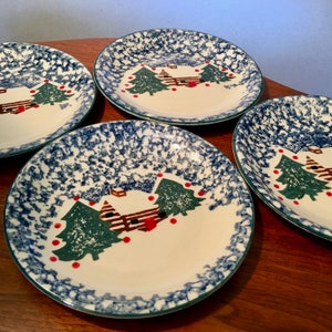 May include: Four white ceramic plates with a Christmas tree design. The plates have a blue speckled border and a green rim. The design features green Christmas trees, a brown fence, and red ornaments. The plates are on a wooden surface.