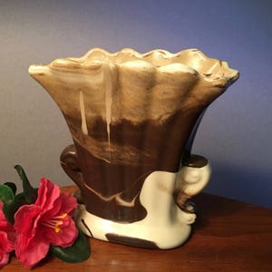 May include: A vintage ceramic vase with a unique, ruffled rim. The vase features a brown and cream marbled design, with a flared shape and two decorative handles. The base is cream-colored. A red flower is visible on the left side.