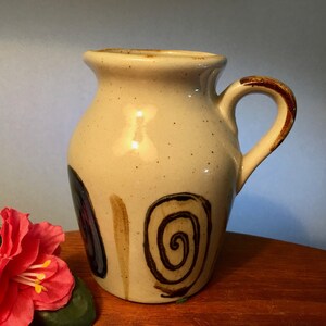 May include: A beige ceramic jug with a brown handle and decorative spiral and stripe designs. The jug has a speckled surface and a slightly flared rim. The design includes dark blue and brown accents.