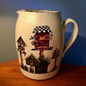 May include: Cream-colored ceramic pitcher with a decorative design of colorful birdhouses. The pitcher has a large handle and a slightly textured rim. The birdhouses are painted in red, green, and black, with birds and floral accents.