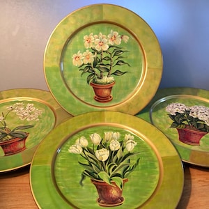 May include: Set of four decorative plates with a green and gold rim. Each plate features a different floral design in a terracotta pot against a green background. The flowers include white orchids, tulips, and hydrangeas.