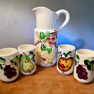 May include: A white ceramic pitcher and four matching tumblers. The pitcher and tumblers are decorated with colorful fruit designs, including grapes, pears, and strawberries. The pitcher has a curved handle and a spout for pouring.