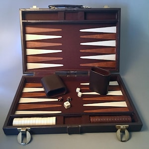 May include: Open backgammon set with a dark brown leather exterior and interior. The playing surface features alternating brown, white, and tan triangles. Includes dice, dice cups, and game pieces. The set is open, revealing the game board.