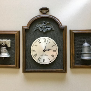 May include: A set of three framed wall decorations. The center piece is a clock with Roman numerals and the text "Seth Thomas". The other two frames contain a Liberty Bell and a pewter tankard. The frames are brown.