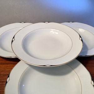 May include: A set of four white porcelain soup bowls. Each bowl has a gold rim and a decorative black and gold accent. The bowls are arranged on a wooden surface.