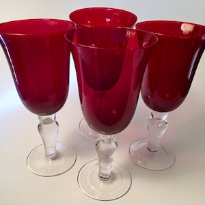 May include: A set of four wine glasses featuring deep red bowls and clear glass stems and circular bases. The glasses have a smooth, rounded silhouette with a wide opening at the top, tapering down to a clear, thick stem and a stable foot.