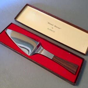 May include: A stainless steel cake server with a wooden handle, presented in a box with red velvet lining. The box lid reads "Sweet Server" by BURNCO.