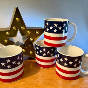 May include: Four ceramic mugs with a patriotic design featuring a blue background with white stars and red and white stripes. A gold star-shaped light fixture is in the background.