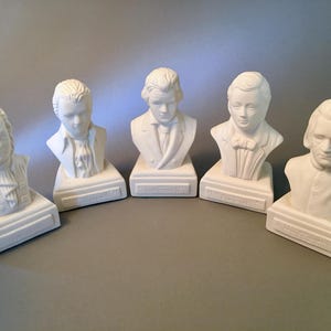 May include: Five white ceramic busts of historical figures. Each bust is mounted on a rectangular base. The busts depict men with various hairstyles and clothing styles, set against a neutral gray background.