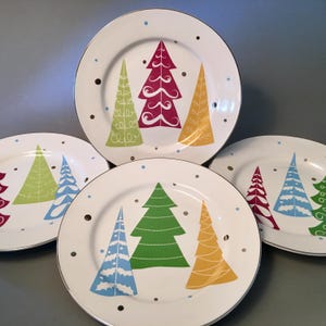 May include: Set of four white ceramic plates with silver rims. Each plate features colorful Christmas tree designs in green, red, yellow, and blue, along with scattered brown and blue dots. A festive holiday theme.