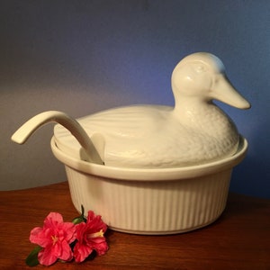May include: A white ceramic duck-shaped serving dish with a lid and spoon. The dish has a ribbed exterior and is on a wooden surface. Two pink flowers are in the foreground.