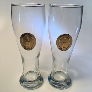 May include: Two clear glass beer glasses with a gold rim and a gold emblem featuring a beer bottle and the words "Corona Extra". The glasses have a curved shape and a wide base.