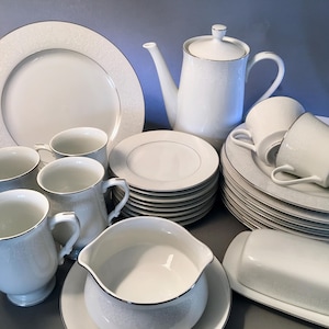 May include: A complete white porcelain dinnerware set with a delicate floral pattern and silver trim. The set includes dinner plates, saucers, cups, mugs, a teapot, a creamer, a gravy boat, and a butter dish.