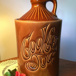 May include: A brown ceramic cookie jar with a lid and handle. The jar is cylindrical with the words "Cookie Jar" embossed on the front in a decorative font. The jar is sitting on a table with a lace doily and pink flowers.
